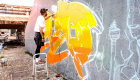 Street artist working on colored graffiti at public space wall - Modern art perform concept of urban guy painting live murales with yellow and orange aerosol color spray - Bright sunflare filter